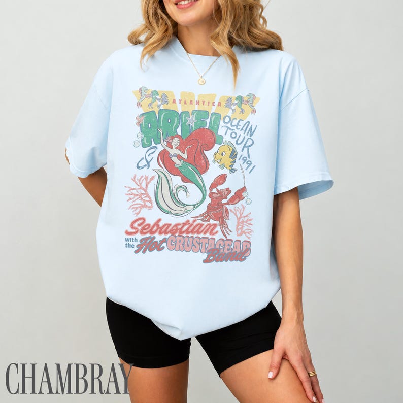 Puede incluir: Una camiseta de manga corta azul claro con un estampado gr&aacute;fico de Ariel, Flounder y Sebasti&aacute;n. El texto dice "Ariel Ocean Tour 1991" y "Sebastian with the Hot Crustacean Band". El dise&ntilde;o incluye ilustraciones de coral y burbujas.