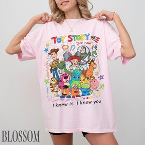 May include: A light pink t-shirt featuring a colourful graphic print of Toy Story characters including Woody, Buzz Lightyear, Jessie, and others. The design includes the text "TOY STORY", "yee haw", and "I knew it I knew you" with scribbled accents.
