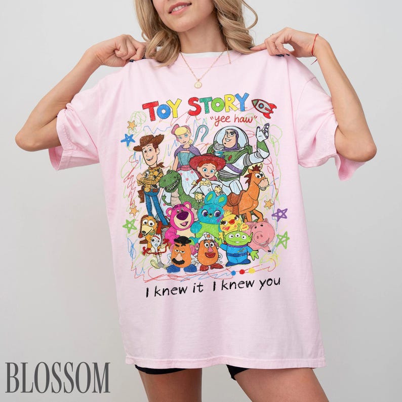 May include: A light pink t-shirt featuring a colourful graphic print of Toy Story characters including Woody, Buzz Lightyear, Jessie, and others. The design includes the text "TOY STORY", "yee haw", and "I knew it I knew you" with scribbled accents.