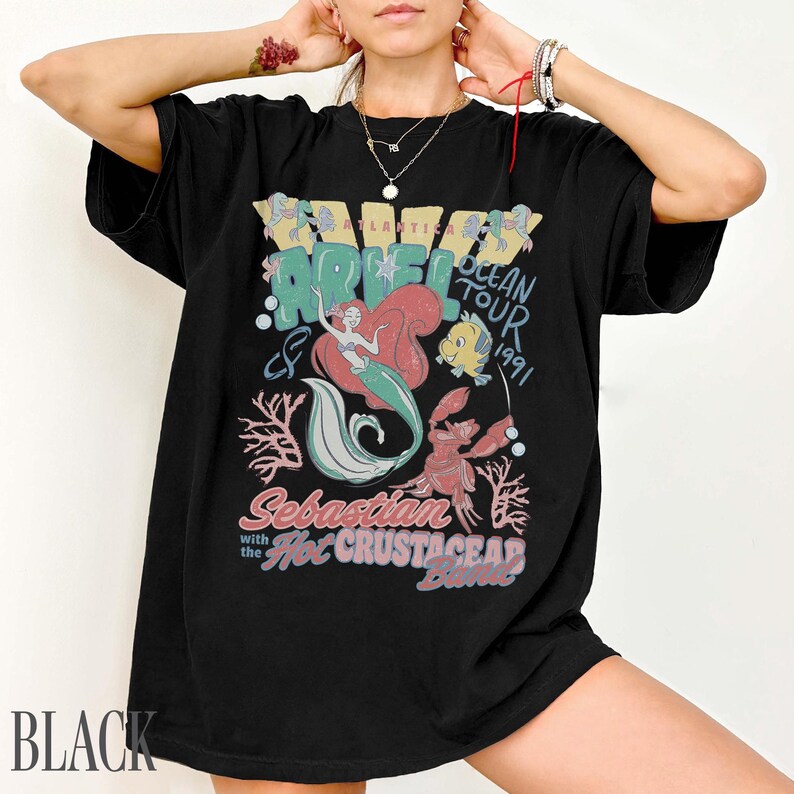 Puede incluir: Una camiseta negra con un estampado gr&aacute;fico de Ariel, Flounder y Sebasti&aacute;n. El texto dice Atlantica Ocean Tour 1991, Sebastian with the Hot Crustacean Band. El dise&ntilde;o incluye ilustraciones de corales y burbujas en tonos rojo, azul y verde.