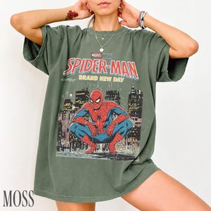 May include: Moss green t-shirt featuring a vintage-style Spider-Man graphic. The design includes the words "MARVEL STUDIOS SPIDER-MAN BRAND NEW DAY" above an illustration of Spider-Man in a city setting. The shirt is a relaxed fit.