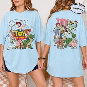 K&ouml;nnte beinhalten: Hellblaues T-Shirt mit einem farbenfrohen Toy Story-Motiv. Auf der Vorderseite sind das Toy Story-Logo und Charaktere wie Woody und Buzz Lightyear abgebildet. Die R&uuml;ckseite des Shirts zeigt das Toy Story-Logo und weitere Charaktere.