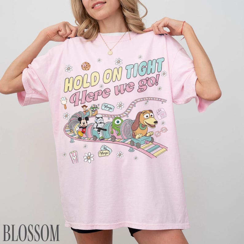 May include: A light pink t-shirt with the text "HOLD ON TIGHT Here we go!" and a colorful graphic of cartoon characters on a roller coaster. The graphic includes Mickey Mouse, a Stormtrooper, and Slinky Dog.