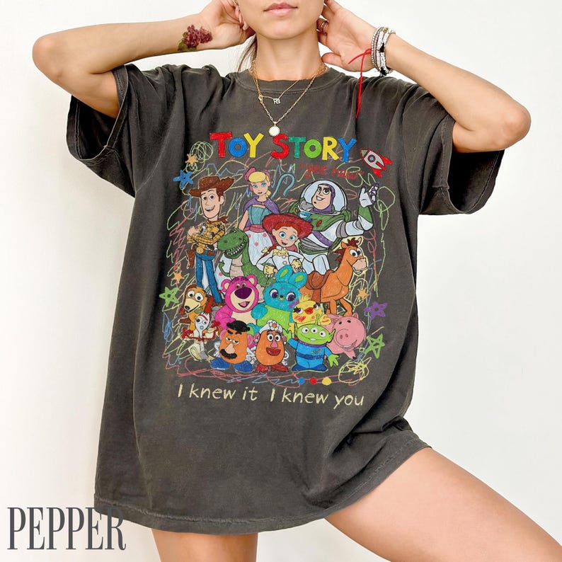 May include: A charcoal grey short-sleeve t-shirt featuring a colourful graphic print of Toy Story characters including Woody, Buzz Lightyear, and Jessie. The design includes scribbled star and circle patterns with the text Toy Story and I knew it I knew you.
