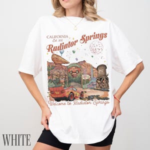 May include: A white short-sleeve t-shirt featuring a graphic print of Radiator Springs with a red race car, a rock formation, a water tower, and the text California Est. 2012 Radiator Springs Welcome to Radiator Springs in brown and red lettering.
