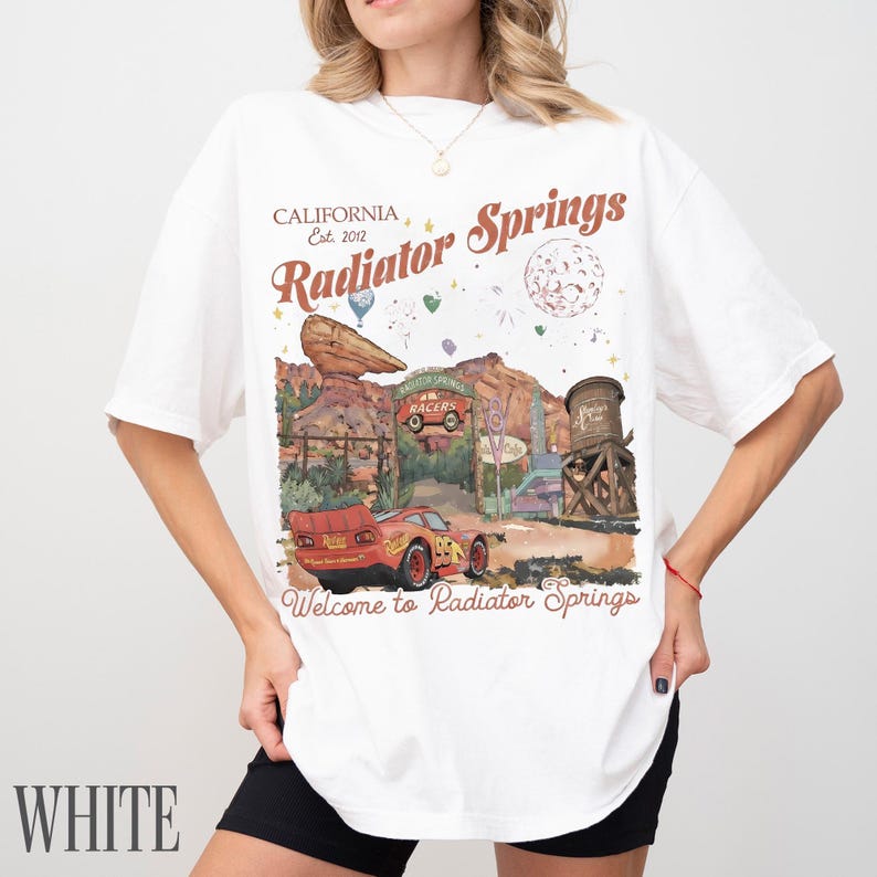 May include: A white short-sleeve t-shirt featuring a graphic print of Radiator Springs with a red race car, a rock formation, a water tower, and the text California Est. 2012 Radiator Springs Welcome to Radiator Springs in brown and red lettering.