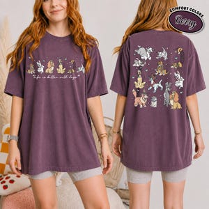 Puede incluir: Camiseta de color baya con un dise&ntilde;o de perros. La parte delantera presenta la frase "life is better with dogs" sobre ilustraciones de perros. La parte trasera de la camiseta muestra varias ilustraciones de perros. La camiseta es de color confort.