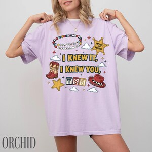 May include: A light purple t-shirt featuring a graphic print with the text I KNEW IT, I KNEW YOU, TS5, INFINITY AND BEYOND, and you've got a friend in me. The design includes a beaded bracelet, a sheriff star, a cowboy boot, a hat, and clouds.