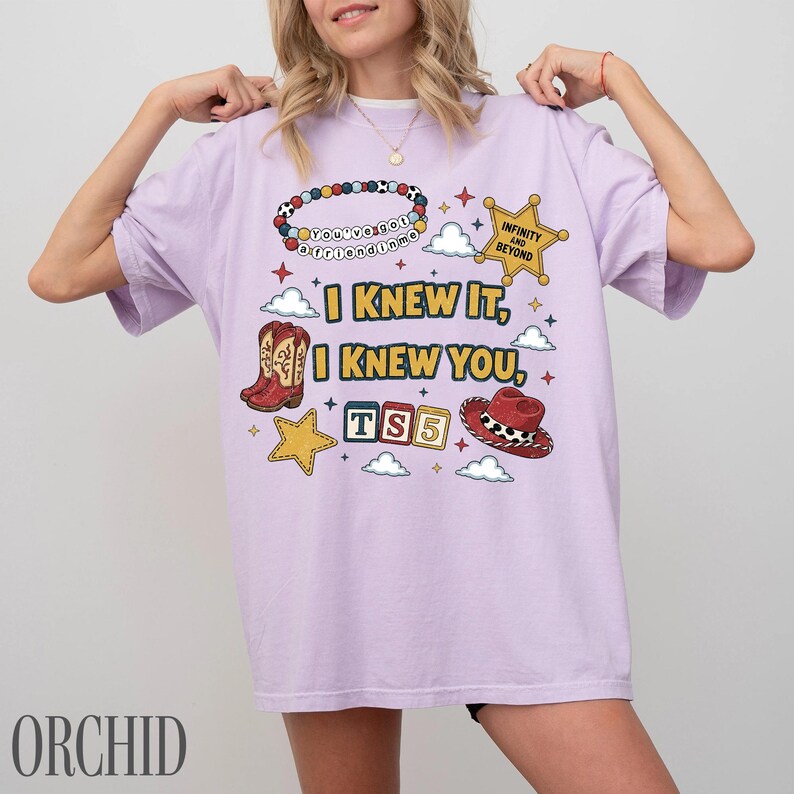 May include: A light purple t-shirt featuring a graphic print with the text I KNEW IT, I KNEW YOU, TS5, INFINITY AND BEYOND, and you've got a friend in me. The design includes a beaded bracelet, a sheriff star, a cowboy boot, a hat, and clouds.