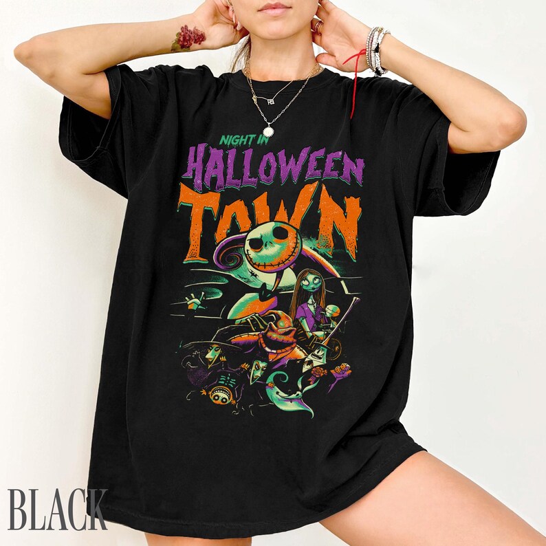 K&ouml;nnte beinhalten: Ein schwarzes Kurzarm-T-Shirt mit einem Grafikdruck und dem Text NIGHT IN HALLOWEEN TOWN in Lila und Orange. Das Design enth&auml;lt Illustrationen verschiedener animierter Figuren, darunter ein Skelett, eine weibliche Gestalt und verschiedene Kreaturen.