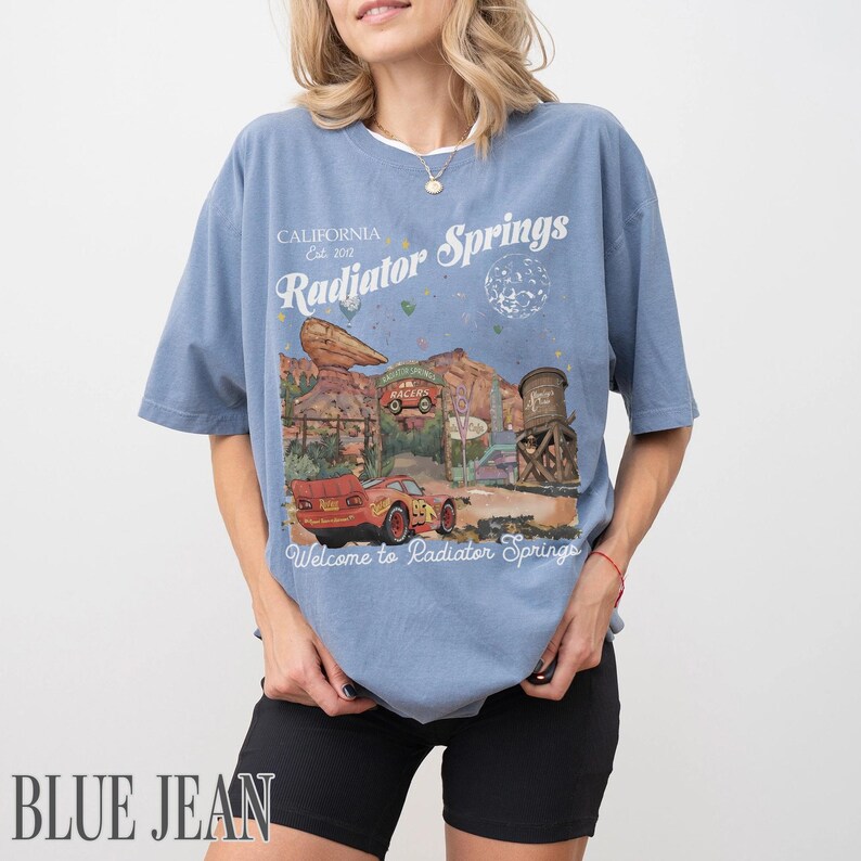 May include: A dusty blue short-sleeve t-shirt featuring a graphic print of a red race car in a desert landscape. The text reads California Est. 2012, Radiator Springs, and Welcome to Radiator Springs, with illustrations of rock formations and a water tower.