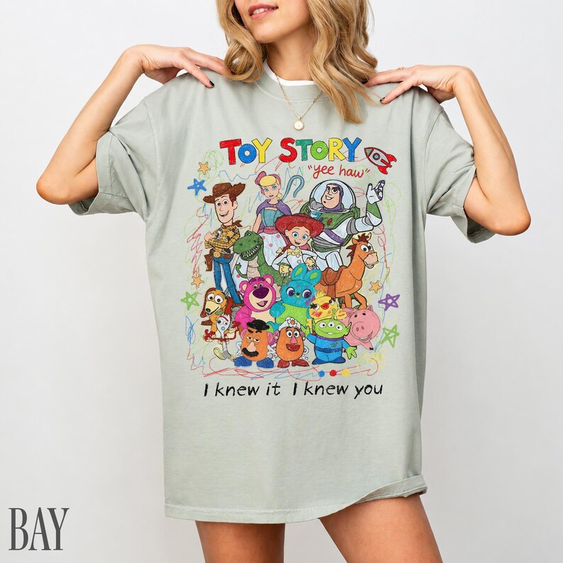 May include: A light grey short-sleeve t-shirt featuring a colourful graphic print of Toy Story characters including Woody, Buzz Lightyear, and Jessie. The text reads "TOY STORY", "yee haw", and "I knew it I knew you" with hand-drawn style stars.