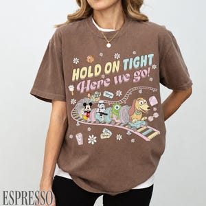May include: Espresso-colored t-shirt with a graphic of a cartoon train carrying Disney characters. The text "HOLD ON TIGHT Here we go!" is printed above the train. The shirt has short sleeves and a crew neck.