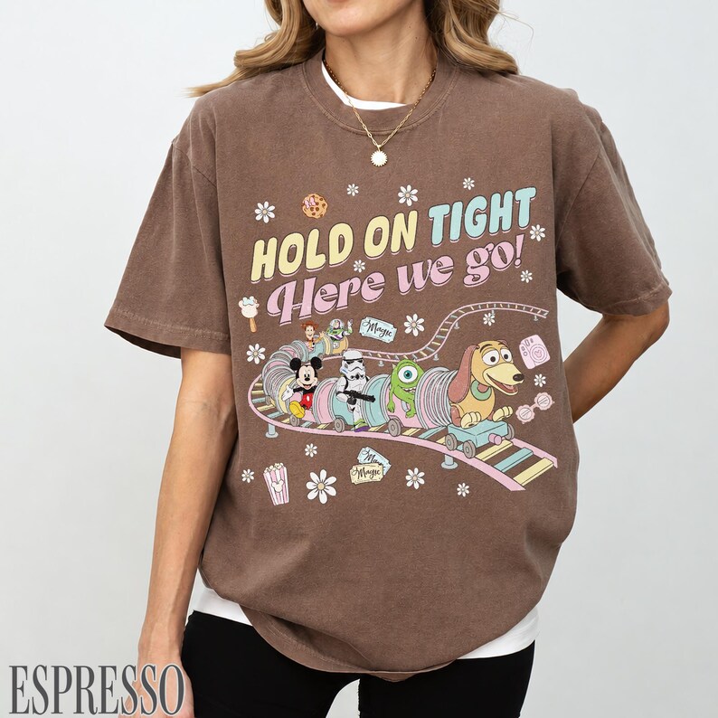 May include: Espresso-colored t-shirt with a graphic of a cartoon train carrying Disney characters. The text "HOLD ON TIGHT Here we go!" is printed above the train. The shirt has short sleeves and a crew neck.