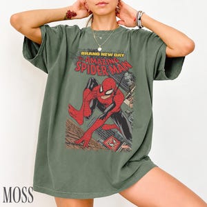 May include: Moss green t-shirt featuring a comic book graphic of Spider-Man in red and black, with the text "BRAND NEW DAY THE AMAZING SPIDER-MAN". The shirt has a vintage, worn look.