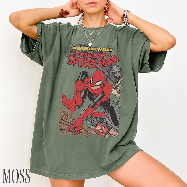 May include: Moss green t-shirt featuring a comic book graphic of Spider-Man in red and black, with the text "BRAND NEW DAY THE AMAZING SPIDER-MAN". The shirt has a vintage, worn look.
