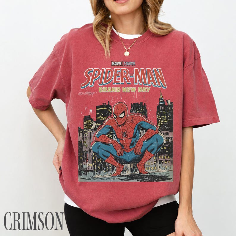 May include: Crimson-colored t-shirt featuring a Marvel Studios Spider-Man graphic. The design includes the words "Spider-Man Brand New Day" and an illustration of Spider-Man in a city setting. The shirt has short sleeves.