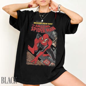 May include: Black t-shirt featuring a vintage comic book graphic of Spider-Man. The design includes the text "BRAND NEW DAY the AMAZING SPIDER-MAN" in red and white. The shirt is a classic crew neck style.