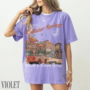 May include: A light purple short-sleeve t-shirt featuring a graphic print of Radiator Springs with a red race car, desert rock formations, and a water tower. The text reads California 2012, Radiator Springs, and Welcome to Radiator Springs.