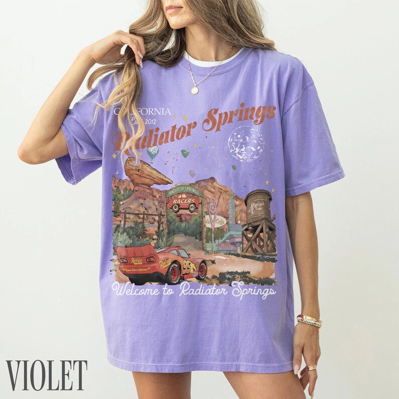 May include: A light purple short-sleeve t-shirt featuring a graphic print of Radiator Springs with a red race car, desert rock formations, and a water tower. The text reads California 2012, Radiator Springs, and Welcome to Radiator Springs.