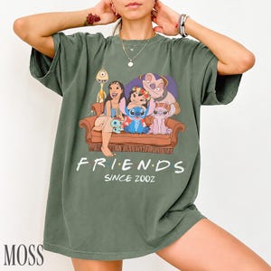 May include: Moss green t-shirt with a graphic of characters from the animated film Lilo & Stitch, seated on a brown couch. The word "FRIENDS" is printed in white, with "SINCE 2002" below. The word "MOSS" is printed in the bottom left corner.