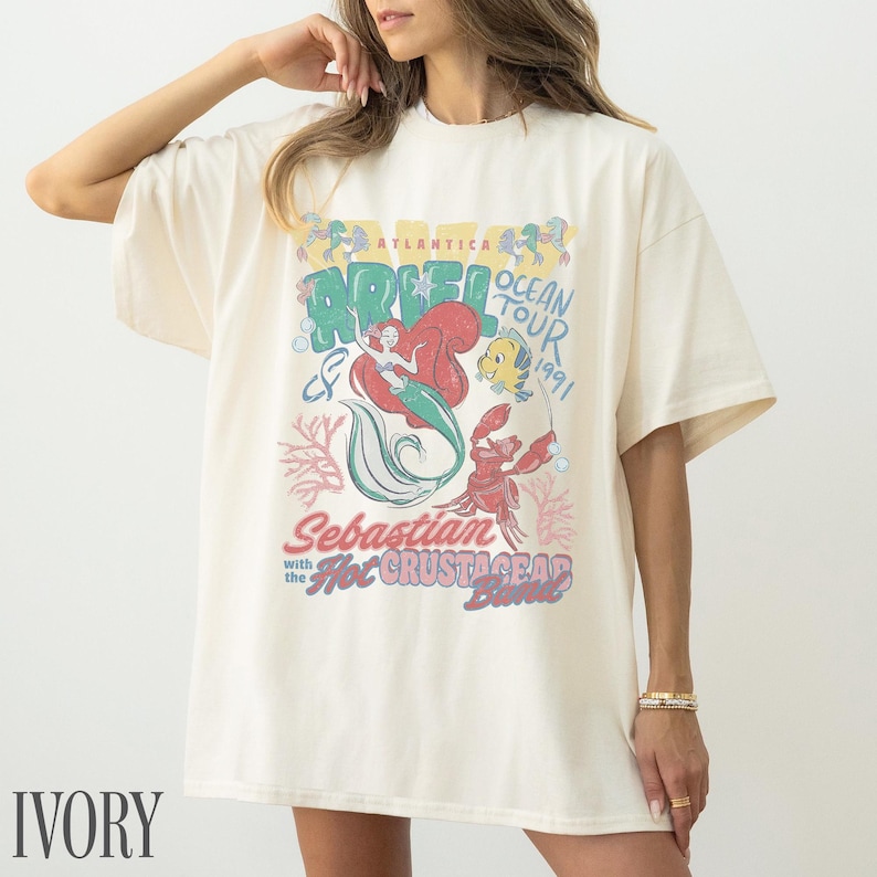 Puede incluir: Una camiseta de manga corta color marfil con un estampado gr&aacute;fico de Ariel, Flounder y Sebasti&aacute;n. El texto dice Atlantica Ocean Tour 1991, Ariel, Sebastian with the Hot Crustacean Band. El dise&ntilde;o incluye ilustraciones de coral y burbujas.