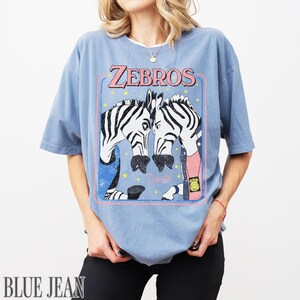 Op de afbeelding: Lichtblauw T-shirt met een afbeelding van twee zebra's en de tekst "ZEBROS" in roze. De zebra's zijn getekend in cartoonstijl, met zwart-witte strepen. Het shirt heeft korte mouwen en een losse pasvorm.