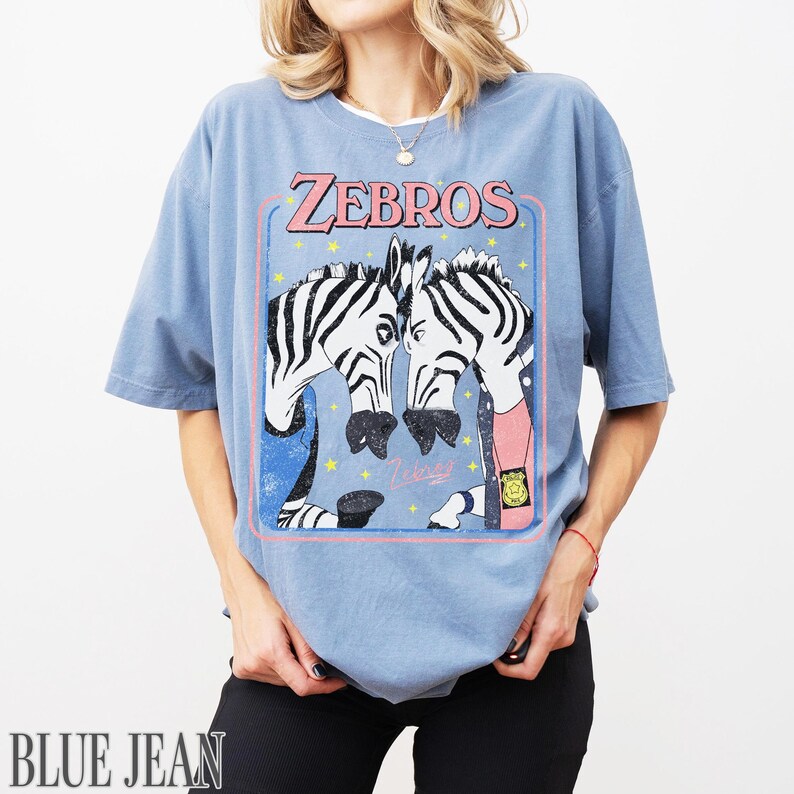 Op de afbeelding: Lichtblauw T-shirt met een afbeelding van twee zebra's en de tekst "ZEBROS" in roze. De zebra's zijn getekend in cartoonstijl, met zwart-witte strepen. Het shirt heeft korte mouwen en een losse pasvorm.