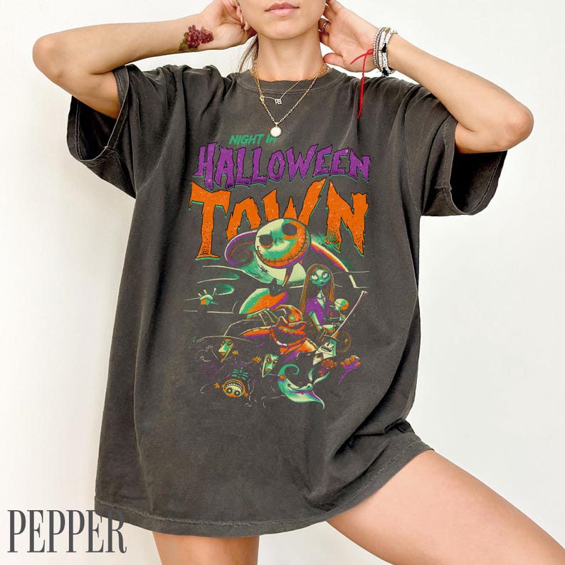 K&ouml;nnte beinhalten: Ein anthrazitfarbenes Kurzarm-T-Shirt mit einem Grafikdruck und dem Text Night in Halloween Town. Das Design enth&auml;lt farbenfrohe Illustrationen verschiedener Figuren, darunter eine skelettartige Gestalt, in den Farben Orange, Lila und Gr&uuml;n.