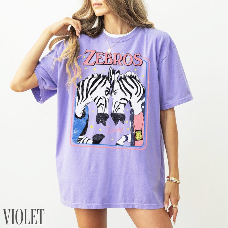 Op de afbeelding: Violetkleurig T-shirt met een afbeelding van twee zebra's die tegenover elkaar staan, omlijst in een roze en blauwe rechthoek. Het woord "ZEBROS" staat in roze boven de zebra's gedrukt. Het woord "VIOLET" staat onderaan het T-shirt.