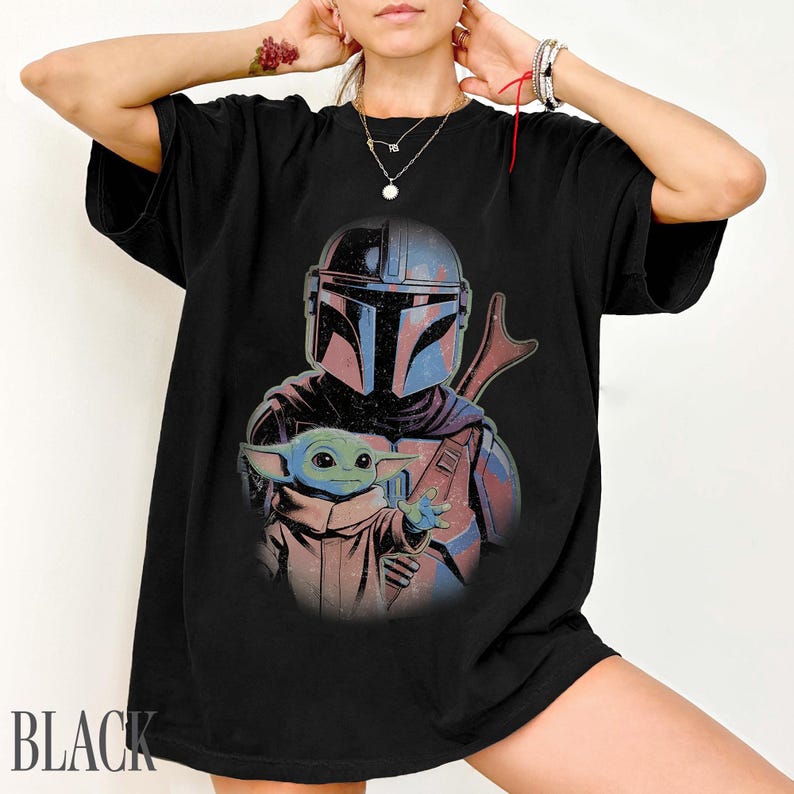 Puede incluir: Una camiseta negra de manga corta con un estampado gr&aacute;fico de un guerrero con casco y armadura oscura detr&aacute;s de una peque&ntilde;a criatura verde de orejas grandes. El dise&ntilde;o utiliza tonos azules, rojos y marrones apagados sobre la tela negra.