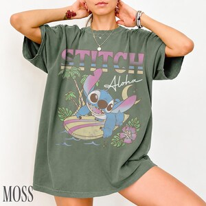 May include: Moss green t-shirt featuring a graphic of Stitch surfing with the word "STITCH" above and "Aloha" below. The design includes palm trees, a crescent moon, and a flower. The shirt is a relaxed fit.