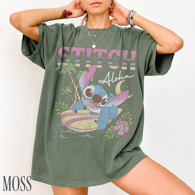 May include: Moss green t-shirt featuring a graphic of Stitch surfing with the word "STITCH" above and "Aloha" below. The design includes palm trees, a crescent moon, and a flower. The shirt is a relaxed fit.