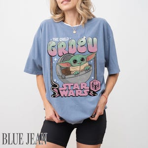 May include: A blue short-sleeve t-shirt featuring a graphic of Grogu from Star Wars inside a floating pod. The text reads THE CHILD GROGU and STAR WARS in a pink and green gradient font. The design includes a small pink helmet icon.