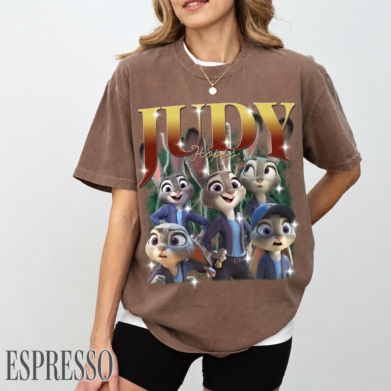 May include: Espresso-colored t-shirt featuring a graphic of Judy Hopps from Zootopia. The design includes the name "JUDY" in gold lettering and images of the character. The shirt is a relaxed fit.