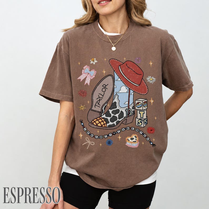 May include: A brown short-sleeve t-shirt featuring a graphic print of a cowboy boot with a cow print pattern, a red cowboy hat, a blue sky background, the name Taylor, the number 13, blocks with letters, and a beaded string with text.