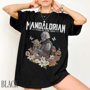 May include: Black t-shirt with the text "MANDALORIAN - THIS IS THE WAY" in white. The design shows the Mandalorian and Grogu with flowers and butterflies. The word "BLACK" is printed at the bottom of the shirt.