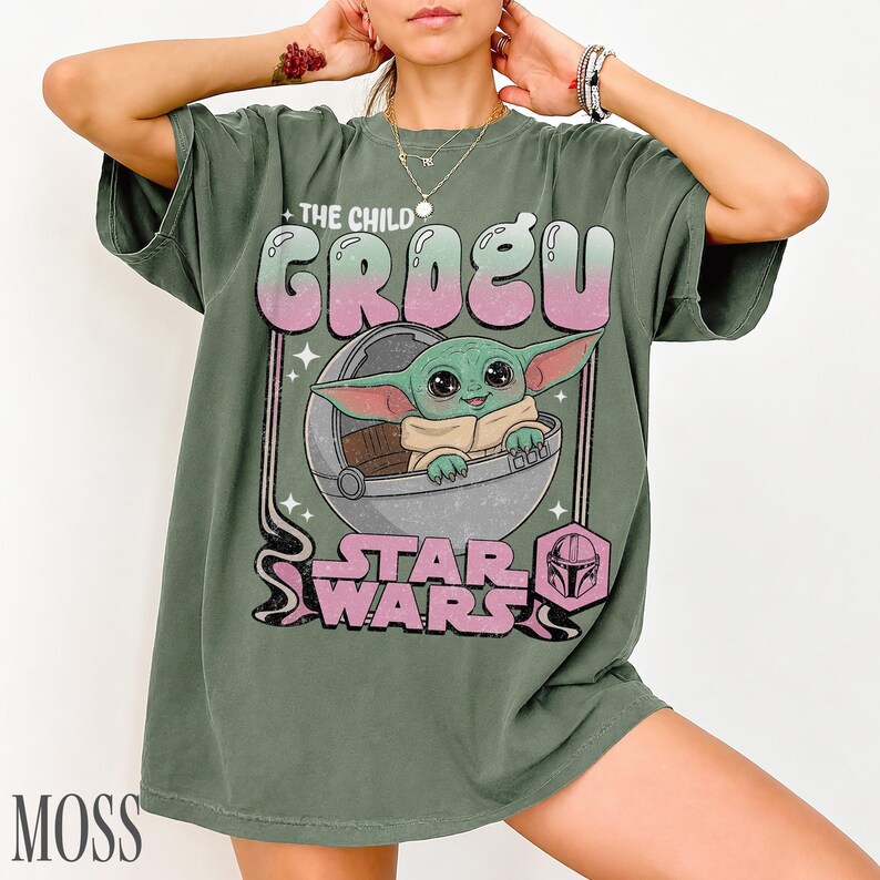May include: A moss green short-sleeve t-shirt featuring a graphic print of Grogu from Star Wars sitting in a floating pod. The text reads The Child Grogu and Star Wars, accompanied by a small helmet icon, all in a faded pink and white color palette.