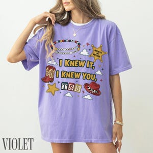 May include: A light purple t-shirt featuring a graphic print with the text "I KNEW IT, I KNEW YOU, TS5", "YOU'VE GOT A FRIEND IN ME", and "INFINITY AND BEYOND". The design includes a beaded bracelet, a sheriff star, cowboy boots, blocks, and a hat.
