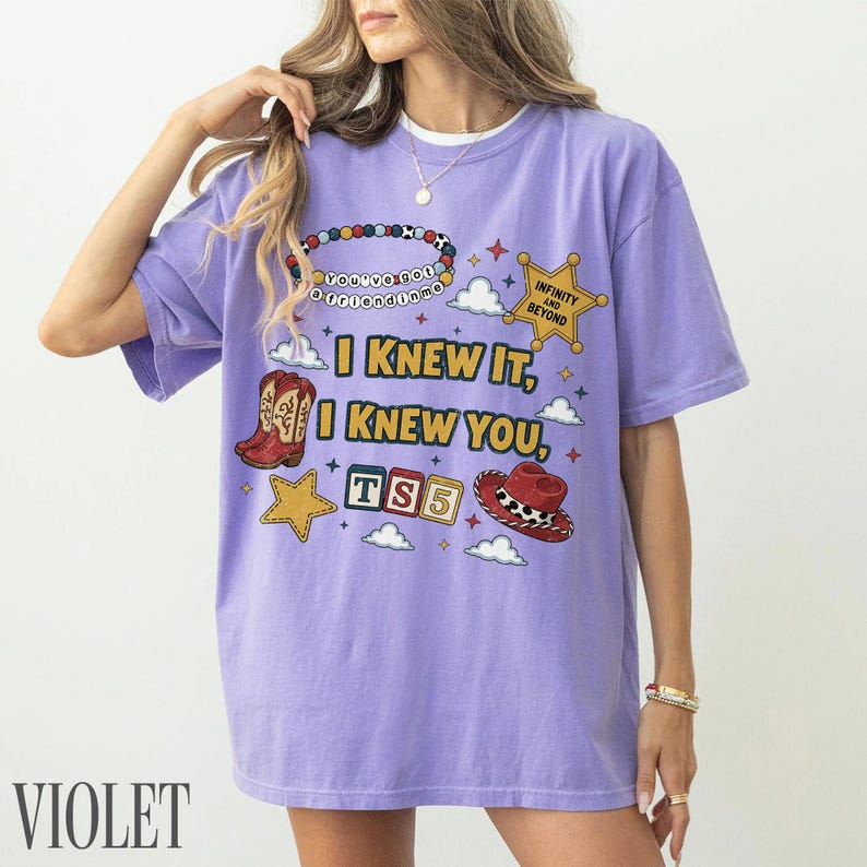 May include: A light purple t-shirt featuring a graphic print with the text "I KNEW IT, I KNEW YOU, TS5", "YOU'VE GOT A FRIEND IN ME", and "INFINITY AND BEYOND". The design includes a beaded bracelet, a sheriff star, cowboy boots, blocks, and a hat.