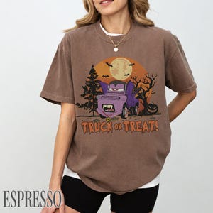 Może przedstawiać: T-shirt w kolorze espresso z grafiką o tematyce Halloween. Projekt zawiera fioletową ciężar&oacute;wkę z kresk&oacute;wek ze skrzydłami nietoperza, pełnię księżyca, nietoperze i napis "TRUCK OR TREAT!". Koszulka ma luźny kr&oacute;j.