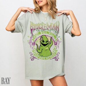 May include: A pale green t-shirt with a graphic of Oogie Boogie from The Nightmare Before Christmas. The design includes the text "BOOGIEMAN" and "WHAT HAVE WE HERE?" in purple and green. The shirt has a loose fit.