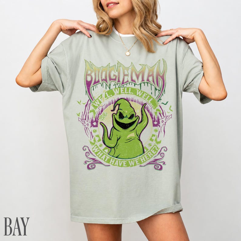 May include: A pale green t-shirt with a graphic of Oogie Boogie from The Nightmare Before Christmas. The design includes the text "BOOGIEMAN" and "WHAT HAVE WE HERE?" in purple and green. The shirt has a loose fit.
