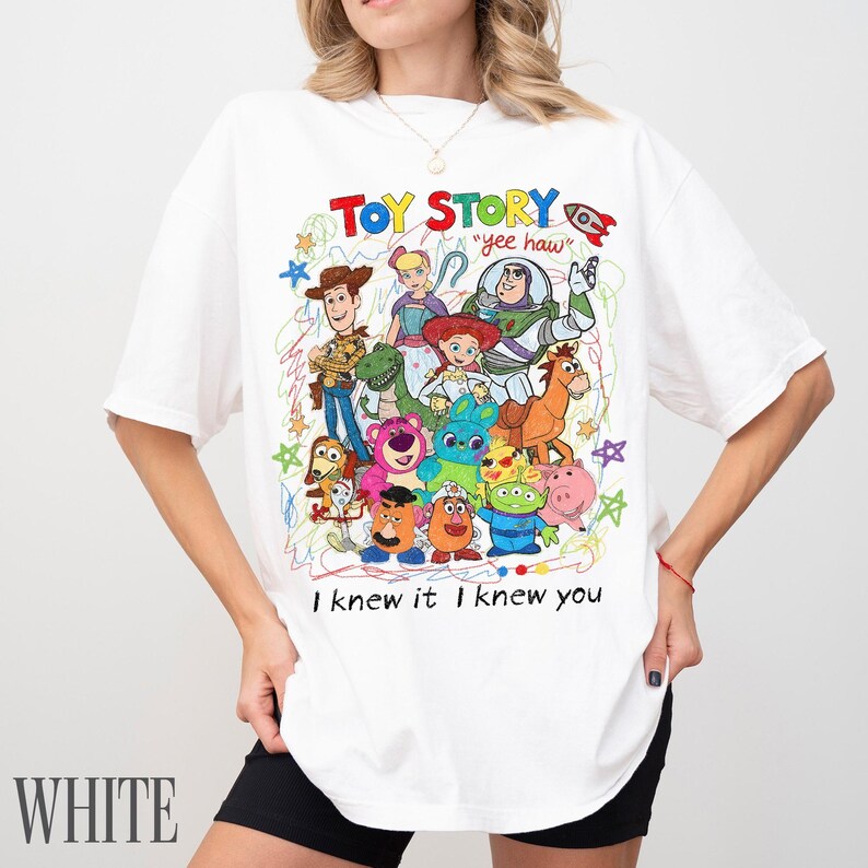 May include: A white t-shirt featuring a colourful graphic print of Toy Story characters including Woody, Buzz Lightyear, Jessie, and others. The design includes the text "TOY STORY", "yee haw", and "I knew it I knew you" surrounded by scribbled star and line art.