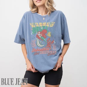 Puede incluir: Una camiseta de manga corta en color azul polvoriento con un estampado gr&aacute;fico de Ariel, Flounder y Sebasti&aacute;n. El texto dice Ariel, Sebastian and the Hot Crustacean Band. El dise&ntilde;o incluye ilustraciones de coral y burbujas en rojo, verde y amarillo.