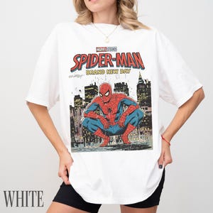 May include: White t-shirt featuring a comic book style graphic of Spider-Man crouching in front of a cityscape. The shirt has the text "MARVEL STUDIOS SPIDER-MAN BRAND NEW DAY". The word "WHITE" is printed at the bottom.
