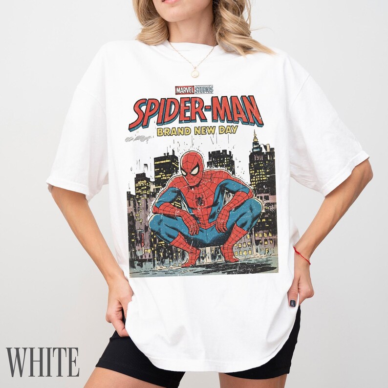 May include: White t-shirt featuring a comic book style graphic of Spider-Man crouching in front of a cityscape. The shirt has the text "MARVEL STUDIOS SPIDER-MAN BRAND NEW DAY". The word "WHITE" is printed at the bottom.