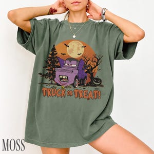 Może przedstawiać: Zielona koszulka z motywem Halloween. Projekt przedstawia fioletową ciężar&oacute;wkę z kłami, pełnię księżyca, nietoperze i napis "TRUCK OR TREAT!". Koszulka ma luźny kr&oacute;j i kr&oacute;tkie rękawy.