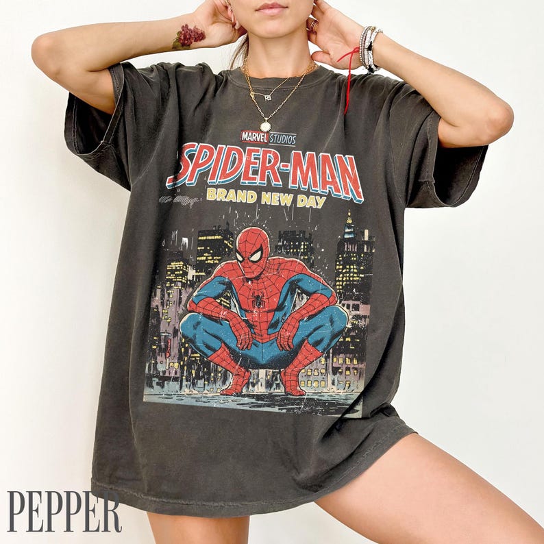 May include: Dark gray t-shirt featuring a Marvel Studios Spider-Man graphic. The design includes Spider-Man in a crouching pose against a cityscape background, with the text "SPIDER-MAN BRAND NEW DAY".