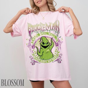 May include: Pale pink t-shirt with a green Oogie Boogie graphic. The design includes the text "BOOGIEMAN" above the character and "WELL WELL WELL... WHAT HAVE WE HERE?" around it. The shirt has a relaxed, oversized fit.